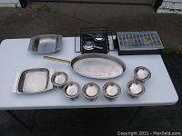 All items in the flambé set laid out on a white table: large oval flambe pan, six small bowls, dual alcohol burner platform, dual warming platform with grate, and two rectangular warming dishes.