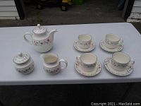 Full view of the white china tea set including teapot, sugar bowl, creamer, and 4 cups with saucers, all decorated with multicolored floral patterns.