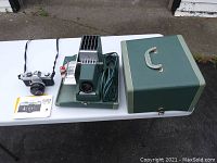 Canon TX 35mm camera with lens, manual, Argus slide projector with power cord, and green carrying case.