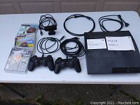 Photo showing the PS3 console, two black DualShock 3 controllers, the PlayStation Eye camera, power and AV cables, user manuals, and the three game cases laid out on a table.
