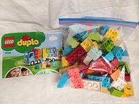 Plastic bag with assorted colorful Lego Duplo pieces including blocks and wheels next to instruction leaflet.