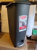 Full view of the tall, slim dark gray Rubbermaid trash can on a cardboard surface, featuring a step-on pedal at the base and an attached label.