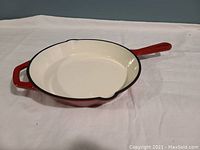 Photo showing a 10-inch red cast iron frying pan with white interior, long handle, and helper handle.