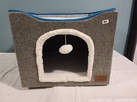 Front angle view showing gray fabric exterior with white plush entrance and hanging white plush toy ball inside the cat cave bed.