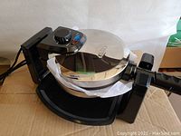 Full side and top view showing the stainless steel rotary waffle maker in open box condition with black base and handle.