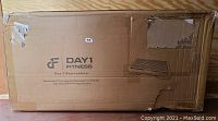 Cardboard box with Day 1 Fitness branding containing aerobic exercise step platform. Box shows shipping wear and damage on one side.