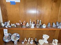 Wide view of multiple figurines and decorative bells arranged on a wooden shelf including cats, birds and various decorative bells.