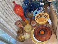 Overview of glass and ceramic ware assortment including tall orange jar, yellow basket, plates, bowls and vases