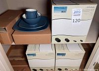 Set of stacked dishes including cups, bowls, salad, and dinner plates in matte blue finish sitting on brown boxes with multiple original boxes seen below.