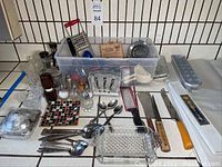 Wide view of assorted kitchen utensils including metal spoons, glass salt shakers, pepper mill, metal grater, napkin holder, and small kitchen tools, displayed on a tiled counter.