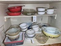 Two shelves of assorted bakeware including red enamel bowls on top left, clear glass pie plates center, white custard bowls top right, various casseroles bottom shelf