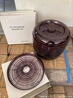 Photo showing the main dark brown translucent electric food dehydrator unit with four stackable trays inside, and one replacement tray outside next to its box labeled 'P917 Food Dehydrator Replacement Tray'. The unit is unplugged and placed on a tile floor.