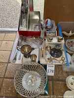 Overview of assorted sterling and silver plated serving pieces arranged on the floor, showing basket, utensils, shot glasses, candlestick and pitcher.