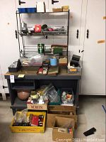 Full view of the small black wood workbench with metal racks and assorted boxes and containers of hardware and tools on and under it.