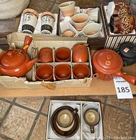 Overview of all stoneware tea sets including teapots, cups, condiment holders, and cups with lids arranged on tile floor.