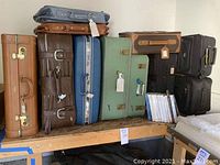 Full view of nine large luggage pieces stacked on wooden table including soft and hard sided suitcases, garment bags, and books.