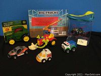 Four Monster Truckin toy vehicles, a John Deere tractor flashlight in pack, and a Big Trucks puzzle pack displayed on blue background.