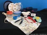 Photo shows the full collection of kitchen and baking items including Tupperware containers, muffin tins, grater, garlic press, apron, oven mitts, measuring cup, and gravy boat on a decorative tablecloth