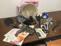 Photo of baking and cooking set with multiple utensils, pans, and cookbooks arranged on table against white wall