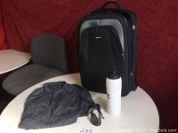 Front view of the black and gray carry-on luggage, gray women's shirt, black sunglasses, and white water bottle on a white table with a gray chair in the background.
