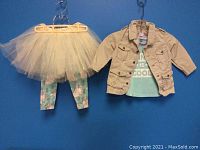 Full view of the child's outfit hanging on two hooks. Shows beige jacket, turquoise tee, leggings with unicorn print, and cream tutu skirt.