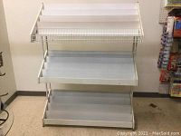 Front view of white metal display rack with three angled shelves against wall and linoleum floor.