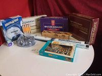Photo of eight games and puzzles on a round table includes Balderdash, Super Quiz, Catch Phrase, Monster Maze, a bag of domino pieces, two puzzle boxes, and a wooden peg game with box.