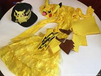 Full view of yellow Pikachu Halloween costume laid out on a flat surface showing dress with hood, tail, and ears along with black Pikachu cap.
