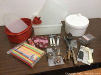 Photo of the full lot showing all items on a wooden table: salad spinner, Tupperware containers, ceramic container, serviette holders, shot glasses, pot holders, utensils and straws.