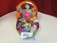 Three small dolls standing inside an orange carrying case filled with various doll clothes.