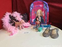Photo showing pink and purple Barbie backpack, Barbie doll sitting in front, pink feather boa, black pillow with pink writing, stuffed unicorn plush toy, and sparkle gray plastic slippers