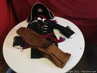 Three-piece children's pirate costume displayed on white round table against red background, showing jacket, pants with boot covers, and pirate hat