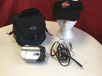 Camcorder with OP/TECH carrying case, power cords, and a black visor cap displayed on a white table.