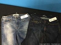 Two pairs of jeans laid side by side on a blue background, one light blue Mavi and one dark wash Hollister, both with tags showing size 27 waist 32 length.