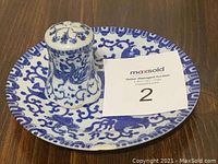 Nippon china salt shaker and matching saucer with blue floral design placed on a surface with a MaxSold auction card showing lot number 2. Salt shaker is damaged as per seller notes.