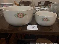 Two white ceramic kitchenware bowls with rose floral designs, different sizes, placed on a wooden table.