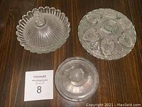 Three assorted glass candy dishes displayed on wood surface. One round divided fruit-shaped section dish, one ribbed scalloped dish with lid, one faceted dish with lid.