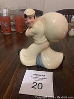 Side view of the vintage ceramic bank figurine depicting a seaman carrying a sack, displayed on a wooden surface with background items.