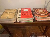 Three stacks of assorted piano sheet music placed on a wooden table, including Beethoven Sonatas on the left, a central collection of various contemporary piano music with music-notes designed red cover on top, and a colorful layered floral cover collection on the right.