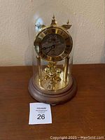 Full view of German anniversary clock under glass dome on a reddish wooden base with visible age wear. Clock face gold-tone with black Roman numerals and ornate center engraving.