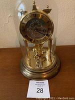 Full view of the brass German anniversary clock inside glass dome with Roman numeral dial and ornate details.