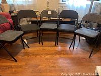 Five folding chairs arranged in a semi-circle on wooden floor, with a lamp and bookshelf in the background. Three chairs have black upholstery, two have gray upholstery.
