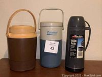 Photo shows three assorted thermoses: a brown insulated jug with beige handle, a blue and silver Coleman thermos, and a black Aladdin vacuum bottle with label.