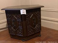 Front view of one ornate wooden end table showing detailed carved wood panels and hinged door with metal handle.