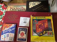 Photo of assorted game boxes and decks including Scrabble in red box, Double Dot Domino playing cards, Bicycle playing cards, wooden dominoes in black box, Connect 4 game, and Chronicles of Narnia box.