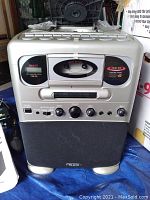 Front view of the silver and black RSQ CGK-90 karaoke machine showing buttons, cassette deck, CD compartment, and microphone inputs.