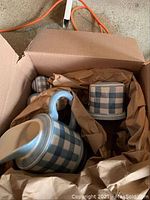 Photo showing box with pottery items wrapped in packing paper: pitcher, napkin holder, salt and pepper shakers with blue and white checkered design.