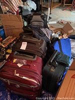 Photo showing multiple luggage pieces stacked, including maroon, black, and gray suitcases, mostly soft-sided with handles and wheels, arranged closely together.