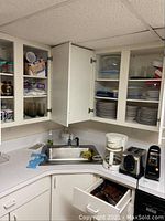 Kitchen corner with white cabinets open showing stacked stoneware plates and bowls, assorted mugs in upper cabinet, kitchen sink with Sunbeam white coffee maker, silver toaster and black/silver electric can opener on counter