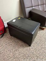 Black faux leather storage ottoman from front, showing closed top and visible wear.